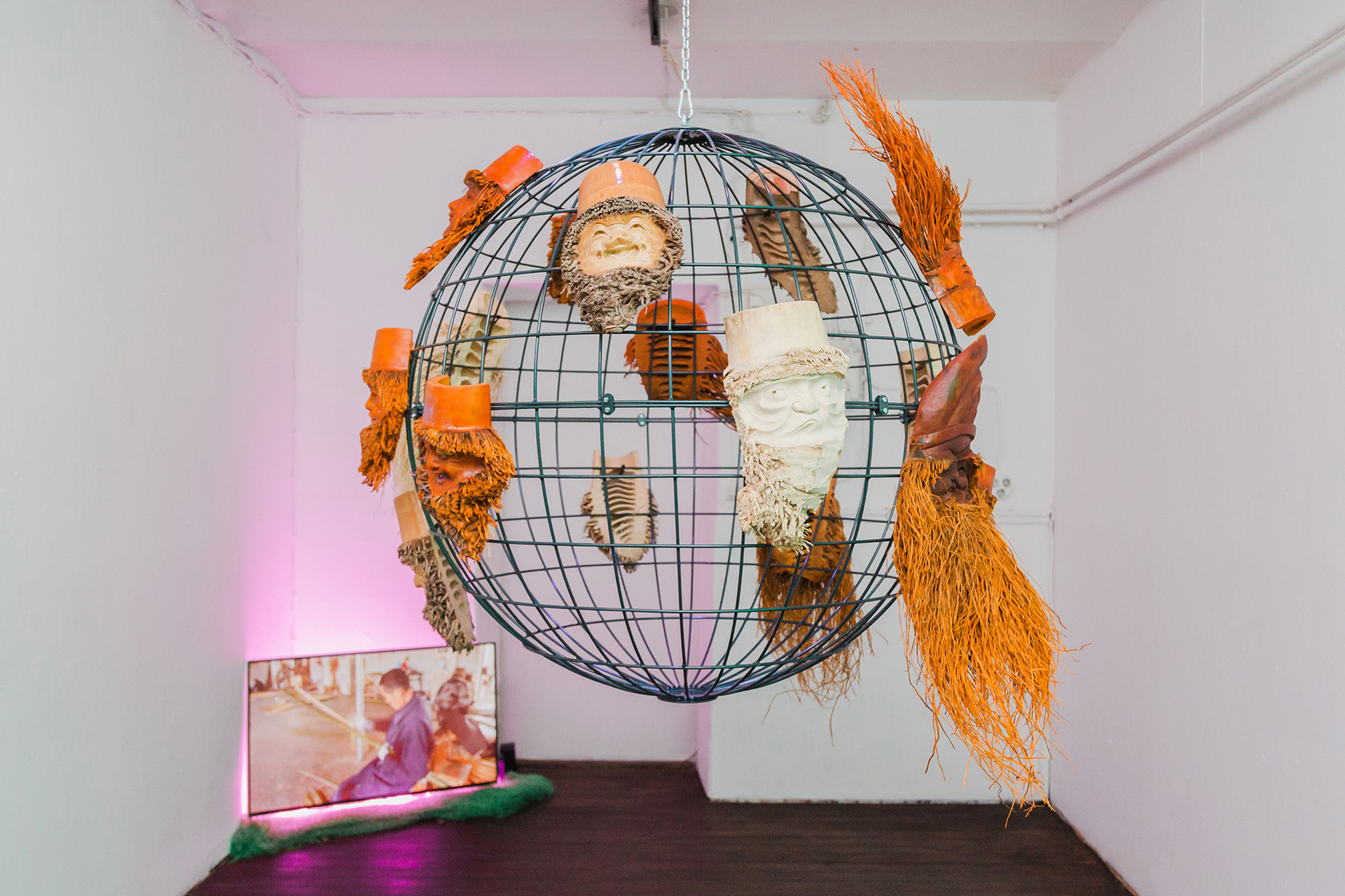 Monika Czyżyk, Bamboo Planet (excerpt / I want to build a rocket), 2020, 4K UHD video, 15 min, colour, stereo sound, pink LED stripe, green mesh, 43” screen, steel sphere, 16 bamboo root masks, zip ties, chain mount, carabiners, 70 cm x Ø 100 cm,
