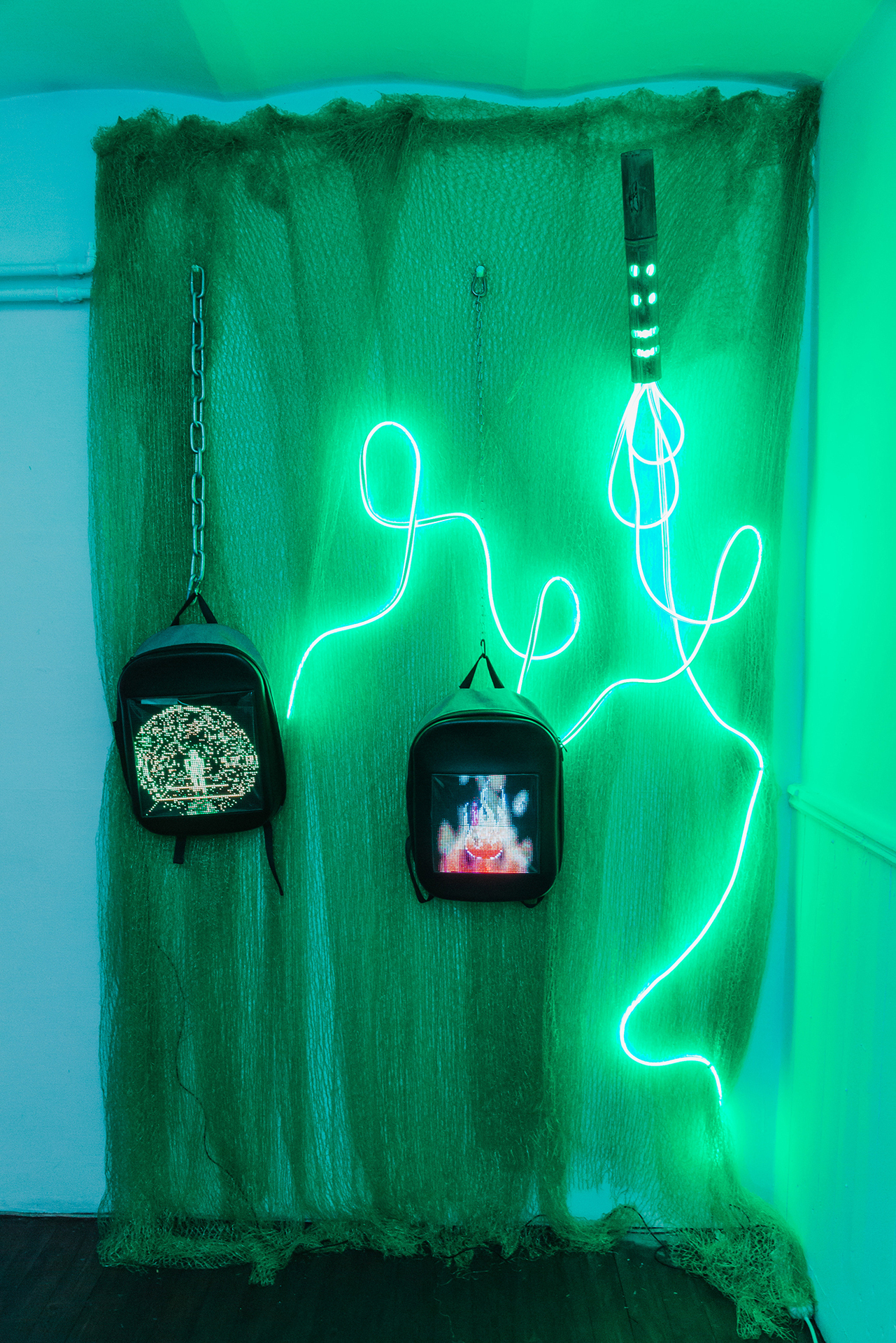 Monika Czyżyk, Green Mesh, 2020, 10” LED screens, backpacks, GIF animations, colour, no sound,  green LED stripe, chains, power bank,  bamboo tube, green mesh, 230 x 15 x 286 cm
