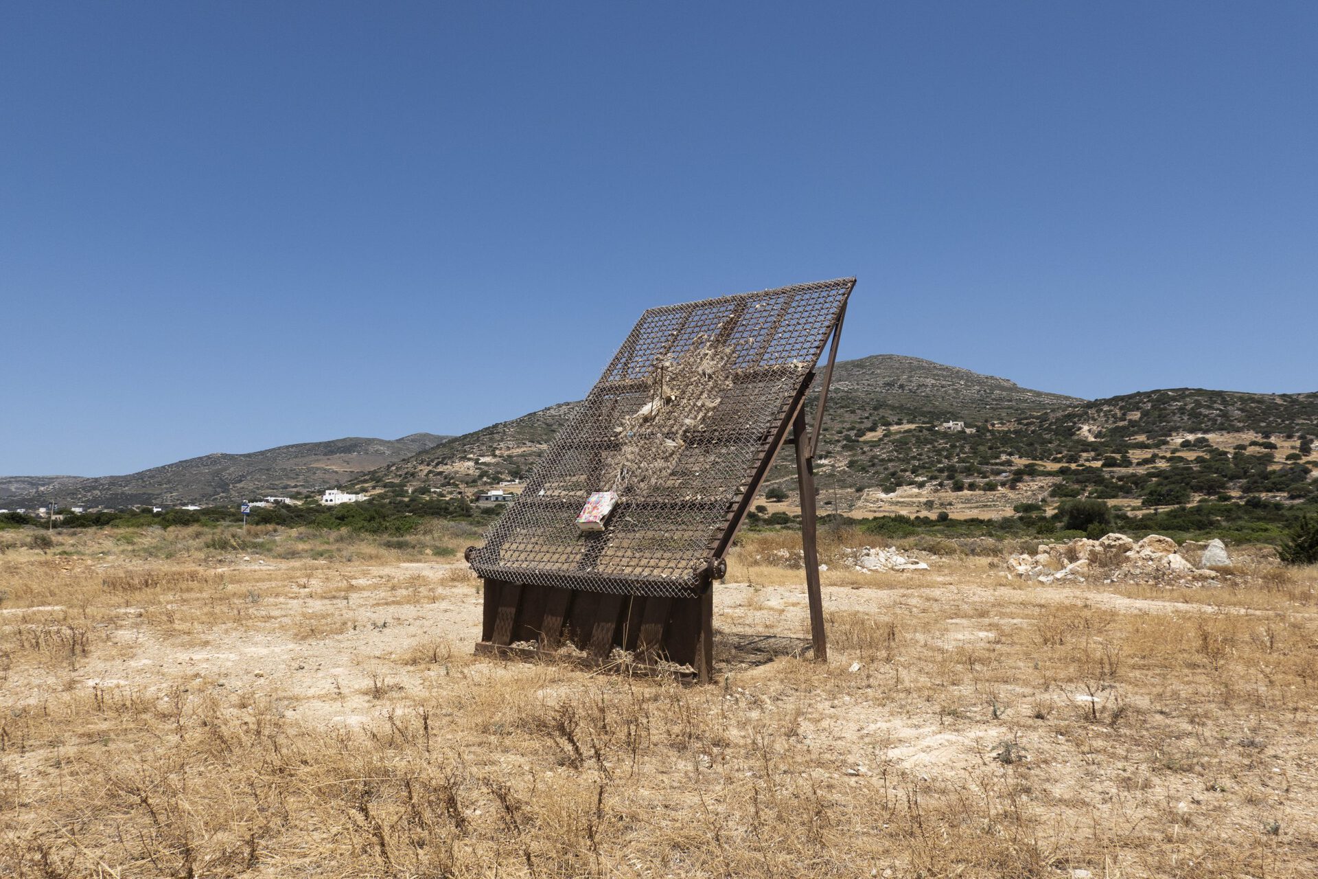 Ishai Shapira Kalter, "Second Stop: Pyrgaki", 2021. Installation view. At the side of the road in a yellow field, a single crate / painting is installed onto an existing, abstract, iron monument. The dating of this monument begins at an unknown time and is now defined as an integral part of the work of artist Ishai Shapira Kalter, who redefined it as a readymade public sculpture.