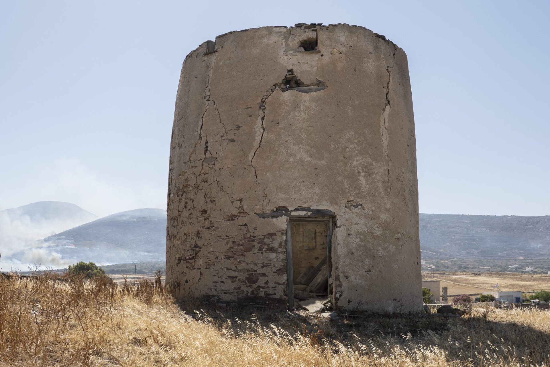 Ishai Shapira Kalter, "Fifth Stop: Lefkes", 2021. Up the mountain, in a ceramics workshop on the outskirts of the Orthodox artists’ village of Lefkes, another crate / painting was supposed to be left by the artist. The humid brown workshop was founded in 1951 and also serves as a souvenir shop for the casual tourist passing by. However, the ceramist did not want to receive the painting as he was occupied looking at th efforts of the Fire Department pilots trying to put out the fire in the mountain across. Therefore, Ishai Shapira Kalter decided to install the work in a nearby destroyed Byzantine windmill down the mountain, overviewing the fire.