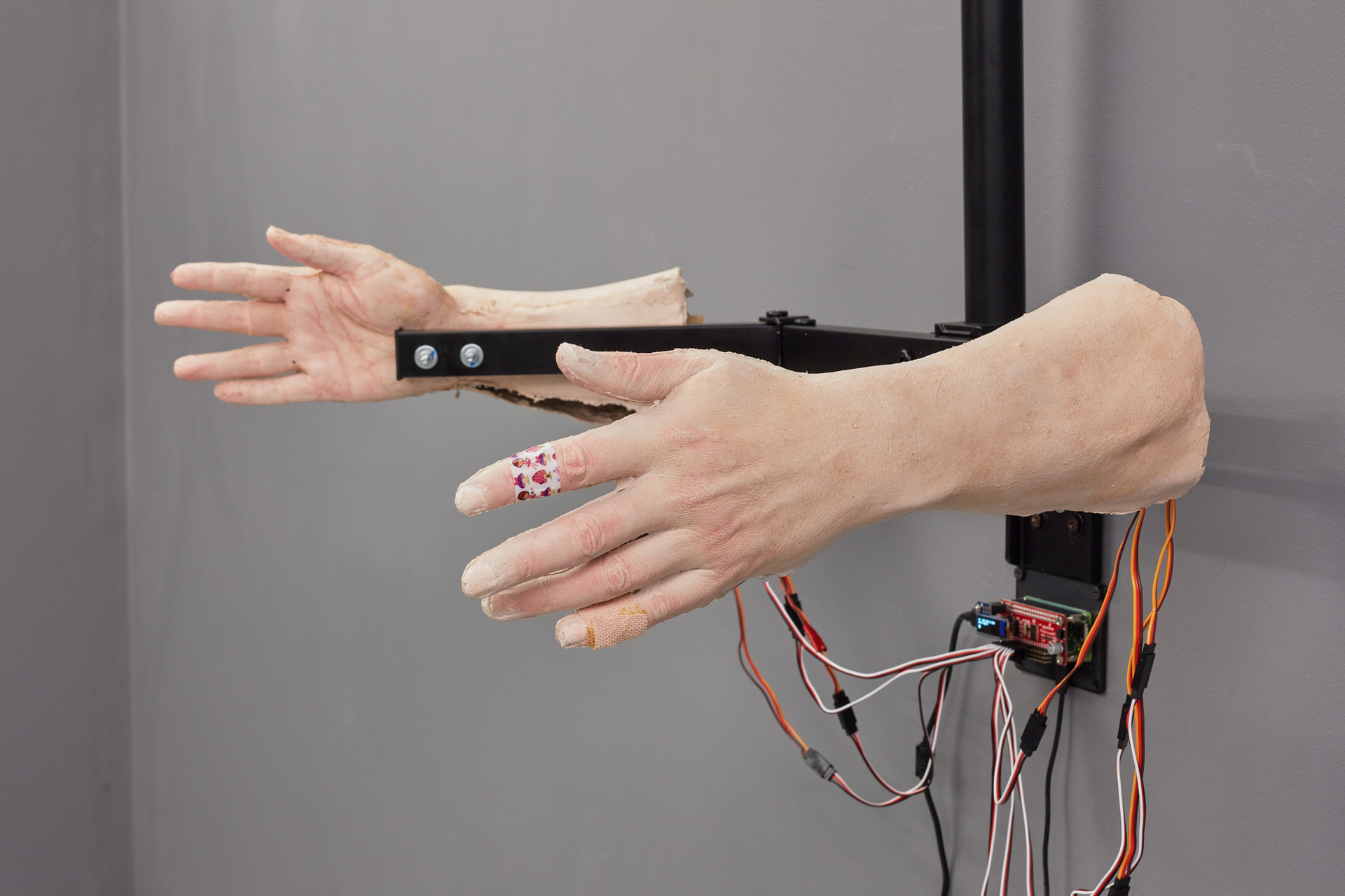 Alex Frost, Passive Aggression 3, 2021, Cast silicon hands, servos, pvc tubing, braided fishing line, wood, Raspberry Pi zero WH, SparkFun Servo pHAT, Adafruit Pio LED & 1A & 3A power supplies, plasters, first aid tape & modified dual monitor stand.