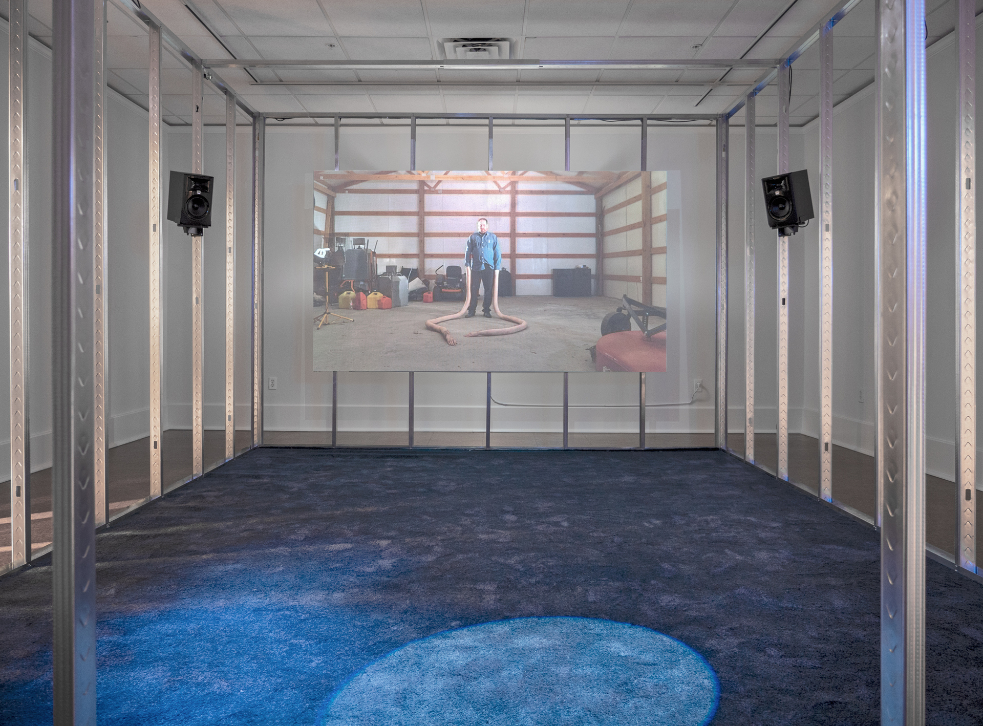 Untitled (blue), 2019; 1080p video projection with sound, blue carpet, drywall, steel studs, door strip curtain, neon signage, speakers, various cables and hardware; installation dimensions variable; video TRT of 7 minutes and 30 seconds, looped