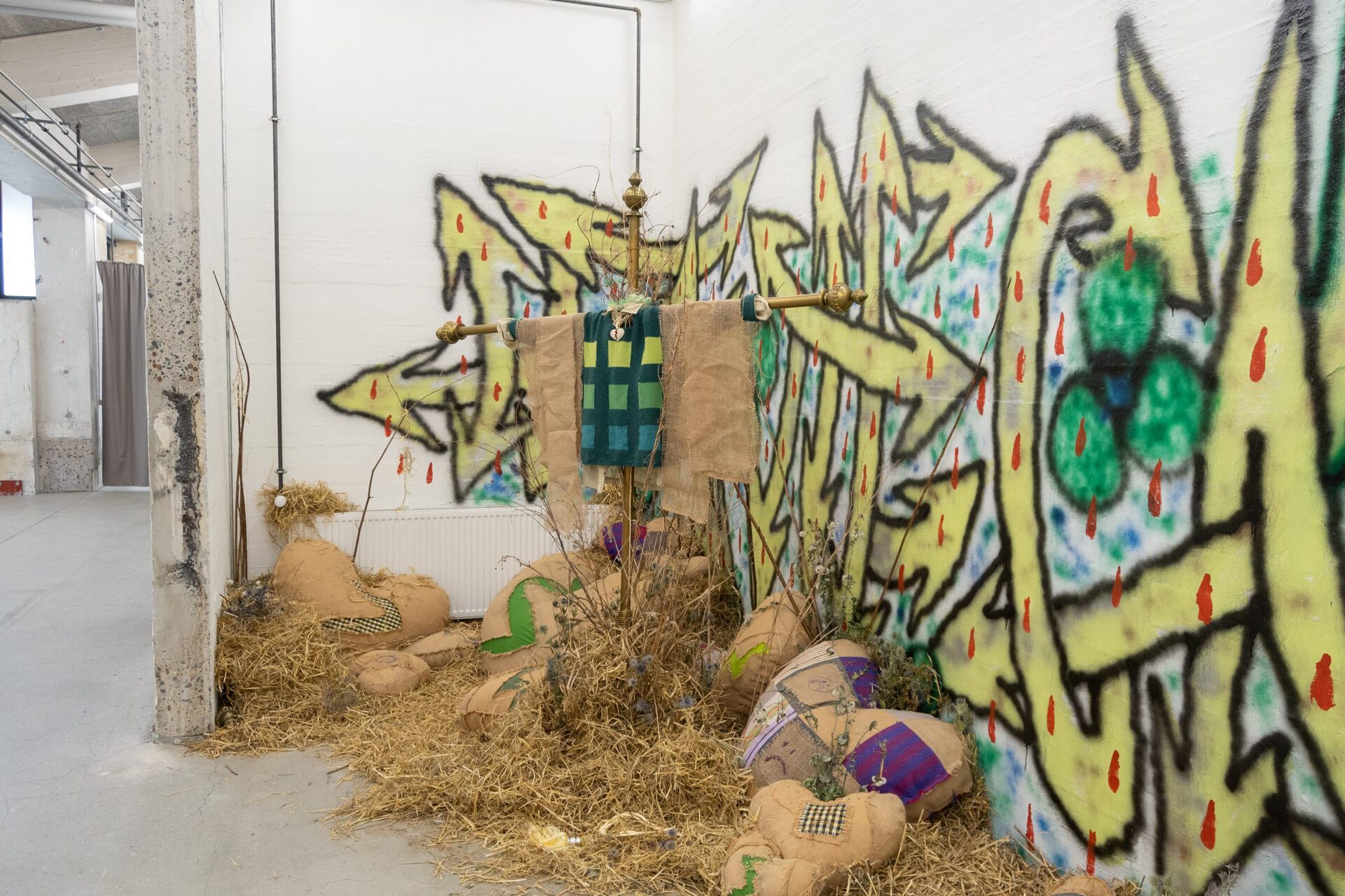 Olivia Rode Hvass, Hate The Game, 2022, Hessian, french curtain rods, heartbreak necklace, hay, roadside-plants, spray paint  (Heart-break pillows made in collaboration with seamstress Fredericke Kellberg – graffiti by anonymous painter)