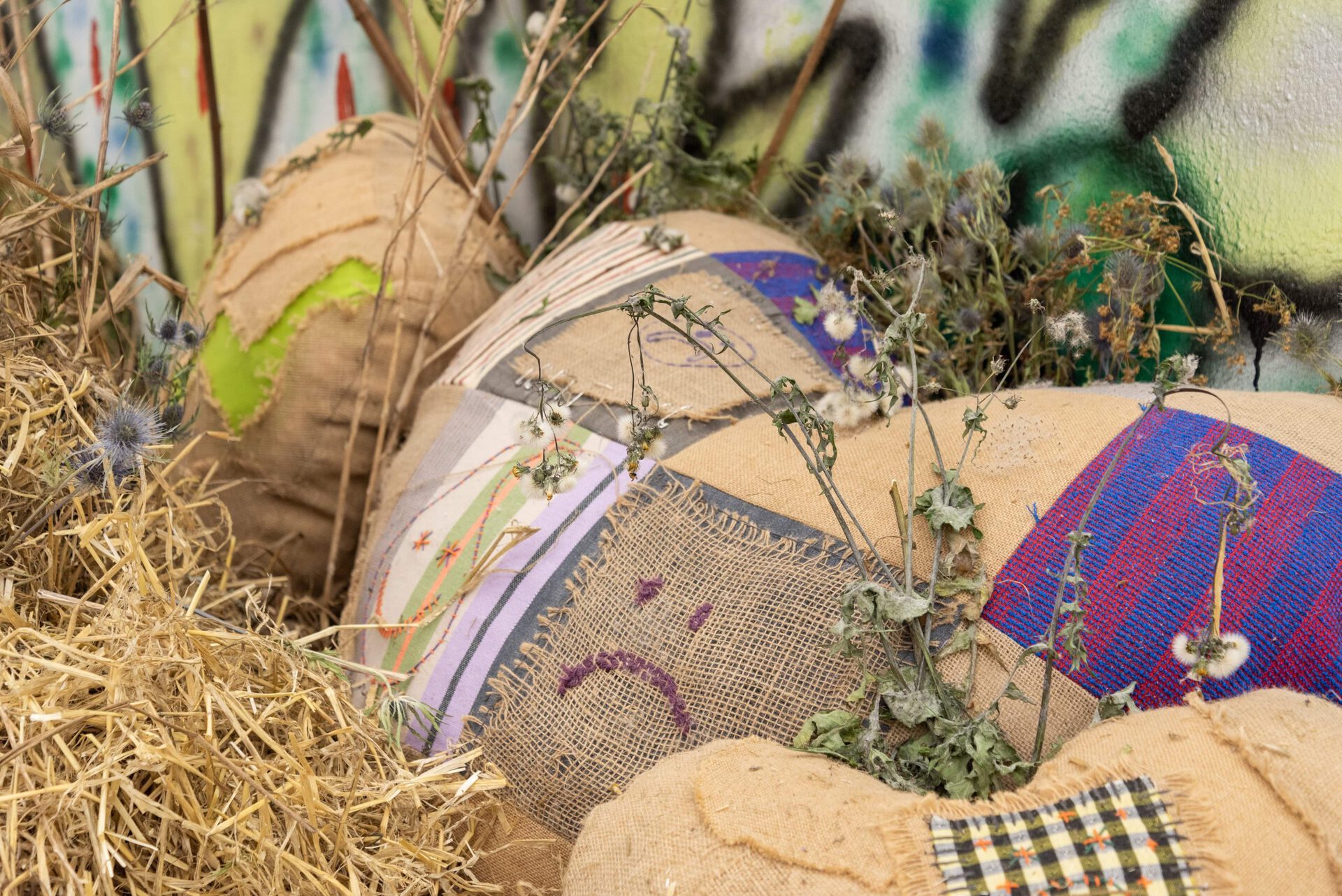 Olivia Rode Hvass, Hate The Game, 2022, Hessian, french curtain rods, heartbreak necklace, hay, roadside-plants, spray paint  (Heart-break pillows made in collaboration with seamstress Fredericke Kellberg – graffiti by anonymous painter)