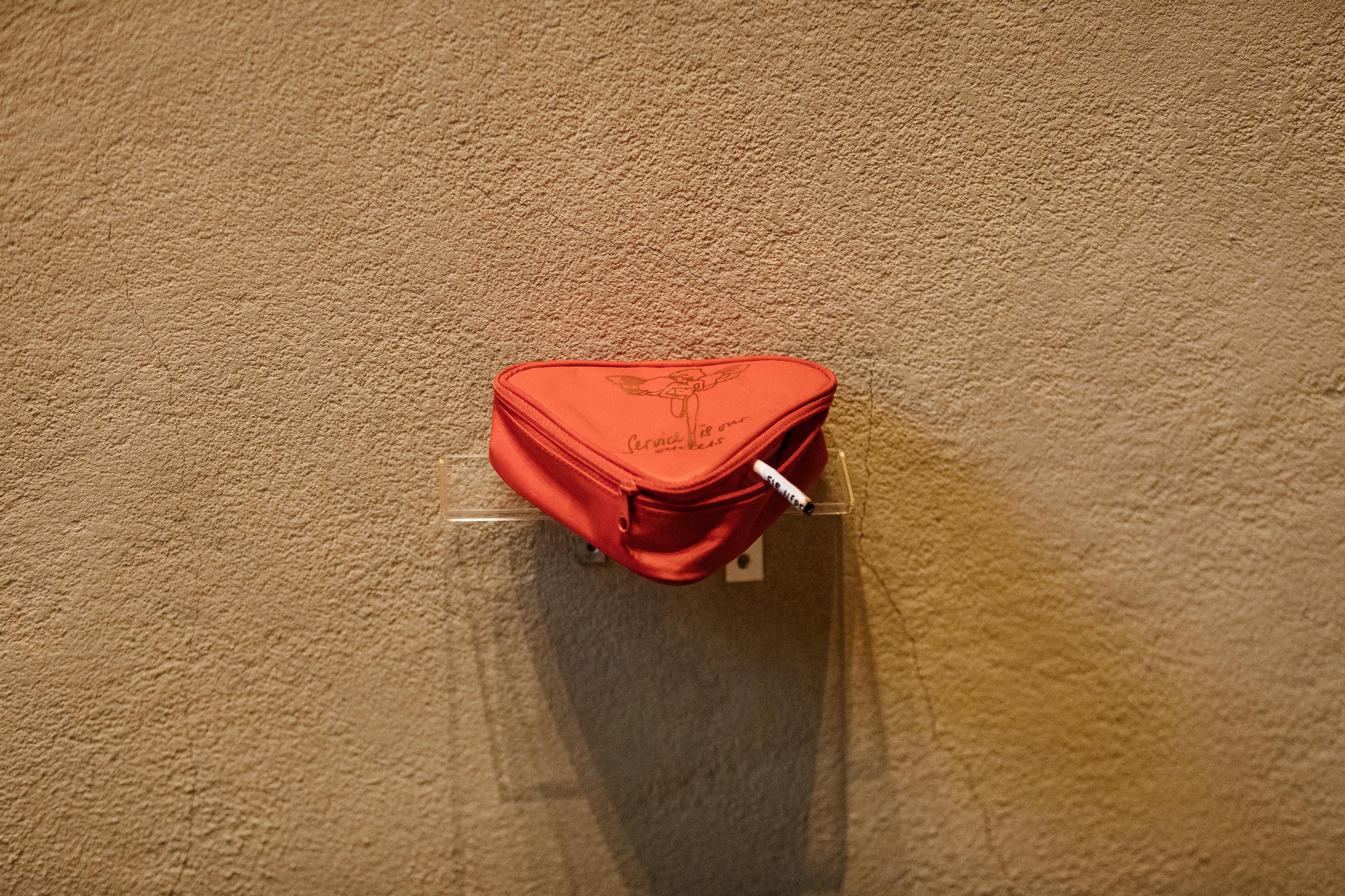 Voyeur, Voyeur - 2022  - material: Lauda Air bag ("Service is our success"), handrolled cigarette with handwritten text (Sir, Alfred), plastic lid, brackets, 17 × 6 × 15 cm