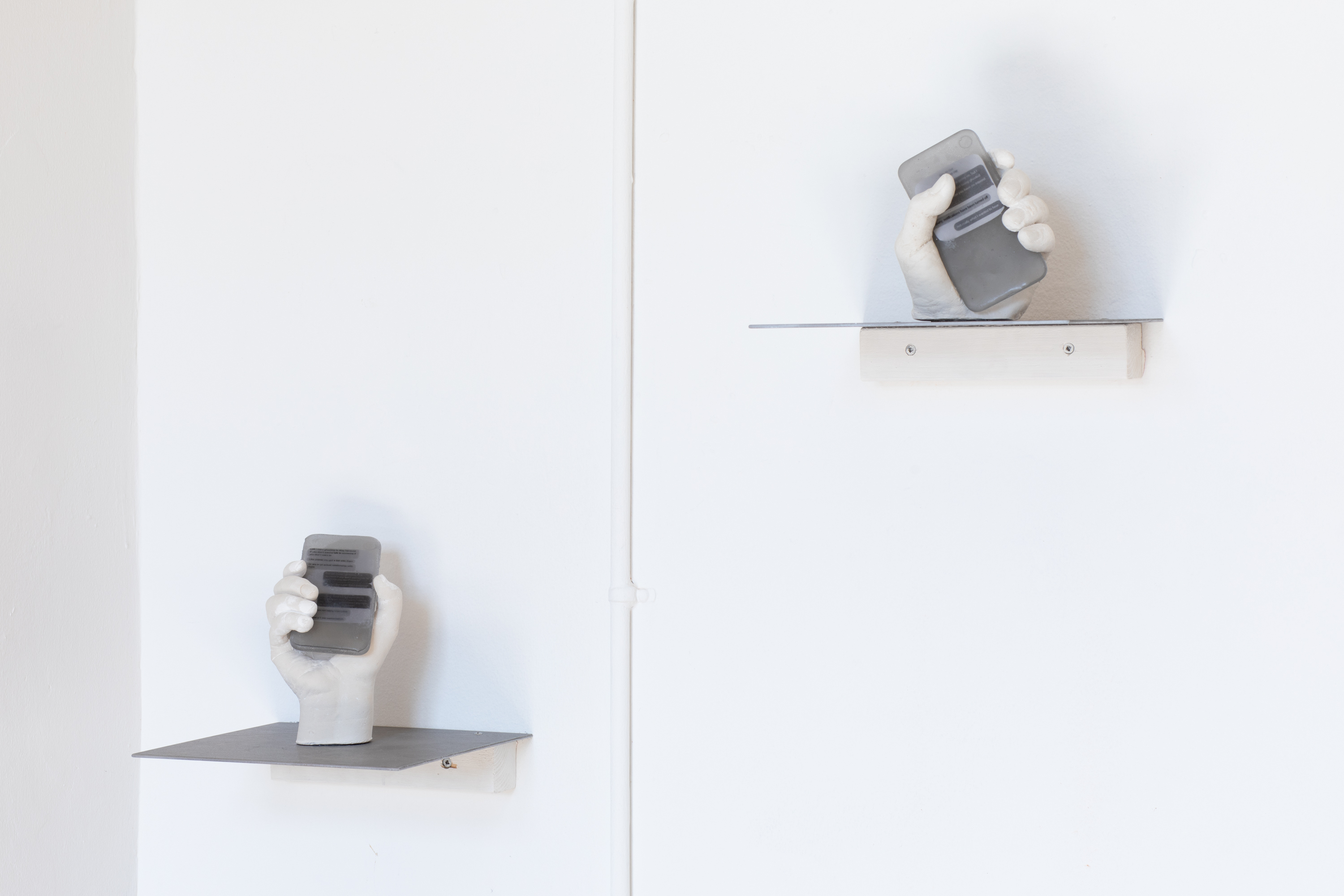Ida Retz Wessberg, âGHOST 11â & âGHOST 12â, 2023 (unique), figure 11: 17,5x13x12 cm, figure 12: 15,5x13x12 cm, isomalt smartphone, plaster hand and steel shelf (25x25 cm), photo: Mads Holm