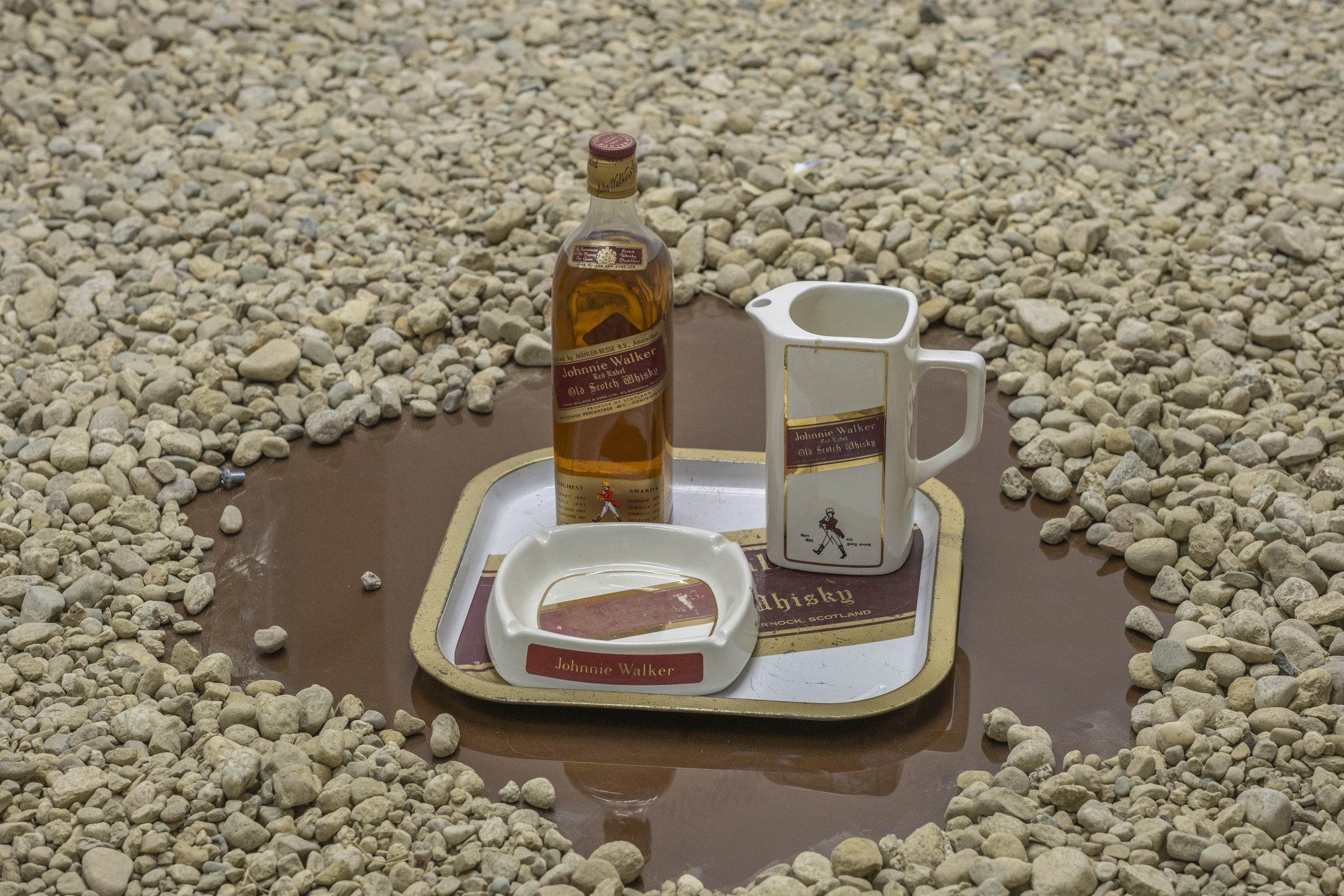 the last good drink was somewhere between 1973 or 1975 (2024) installation plexiglass gravel with found objects: Johnny Walker red label bottle circa 1973, metal tray, ceramic ashtray and pitcher size variable