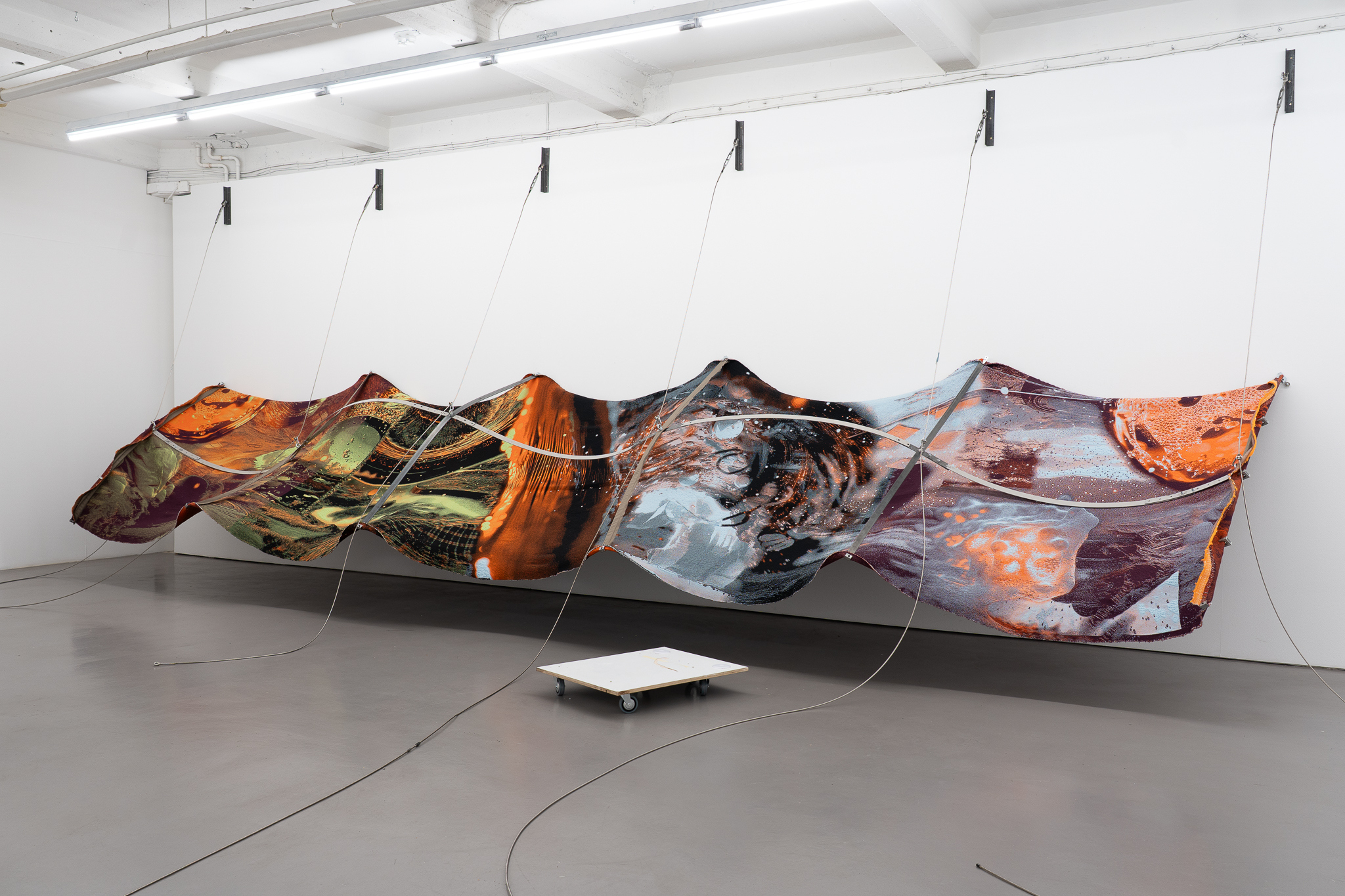 Nicola Arthen - Reading A Reflection (2025); Collage from generated images, machine-knitted wool, cotton and filling yarn, steel rig, C-prints, wooden carts with engravings, work pants, lights, sensors (760x160x145cm), courtesy of the artist and Platform