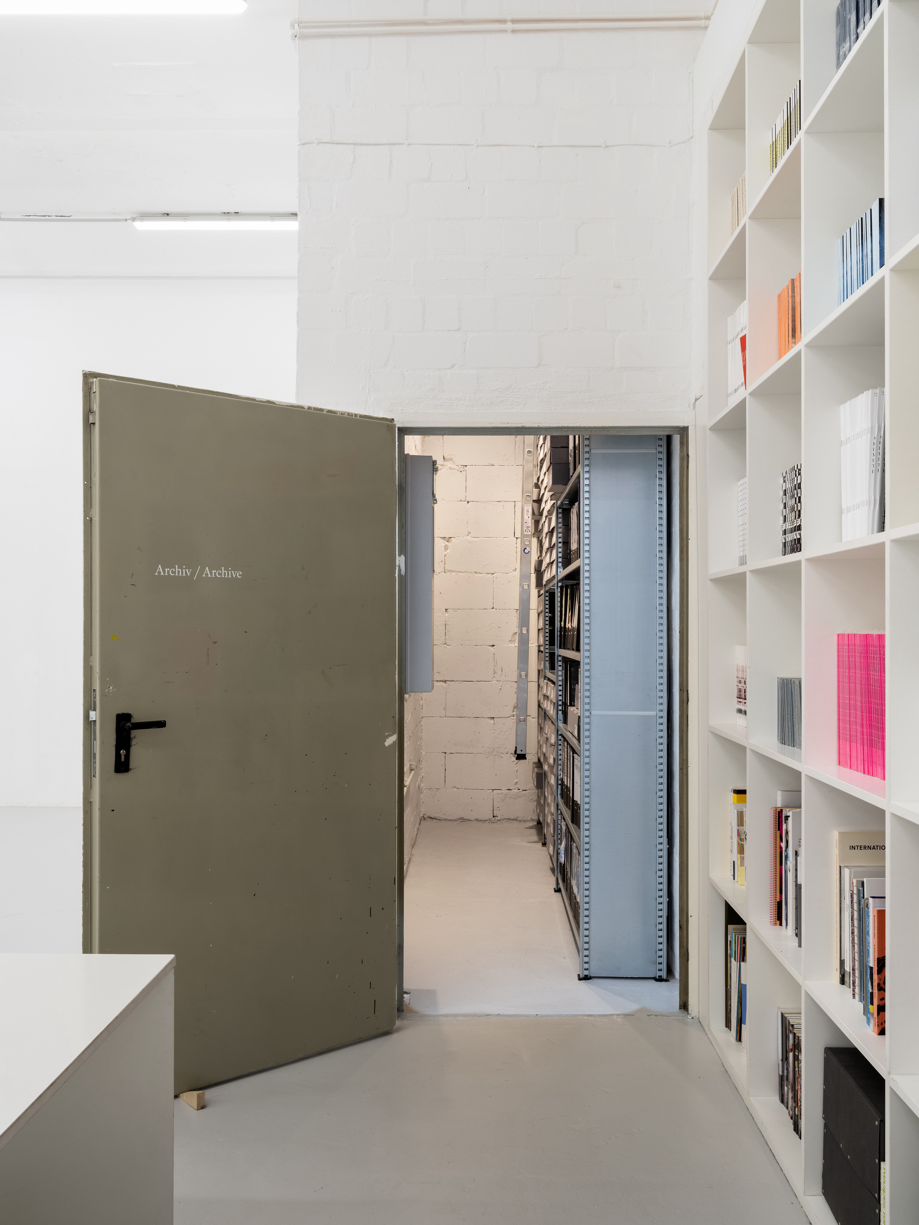 Exhibition view (Main space, Archive), Archiv / Archive Shelving system, lighting system, shelf ladder, wall paint, floor paint, archive boxes with adhesive labels, vinyl lettering, 350x113x391cm, 2025, Photography: Fred Dott