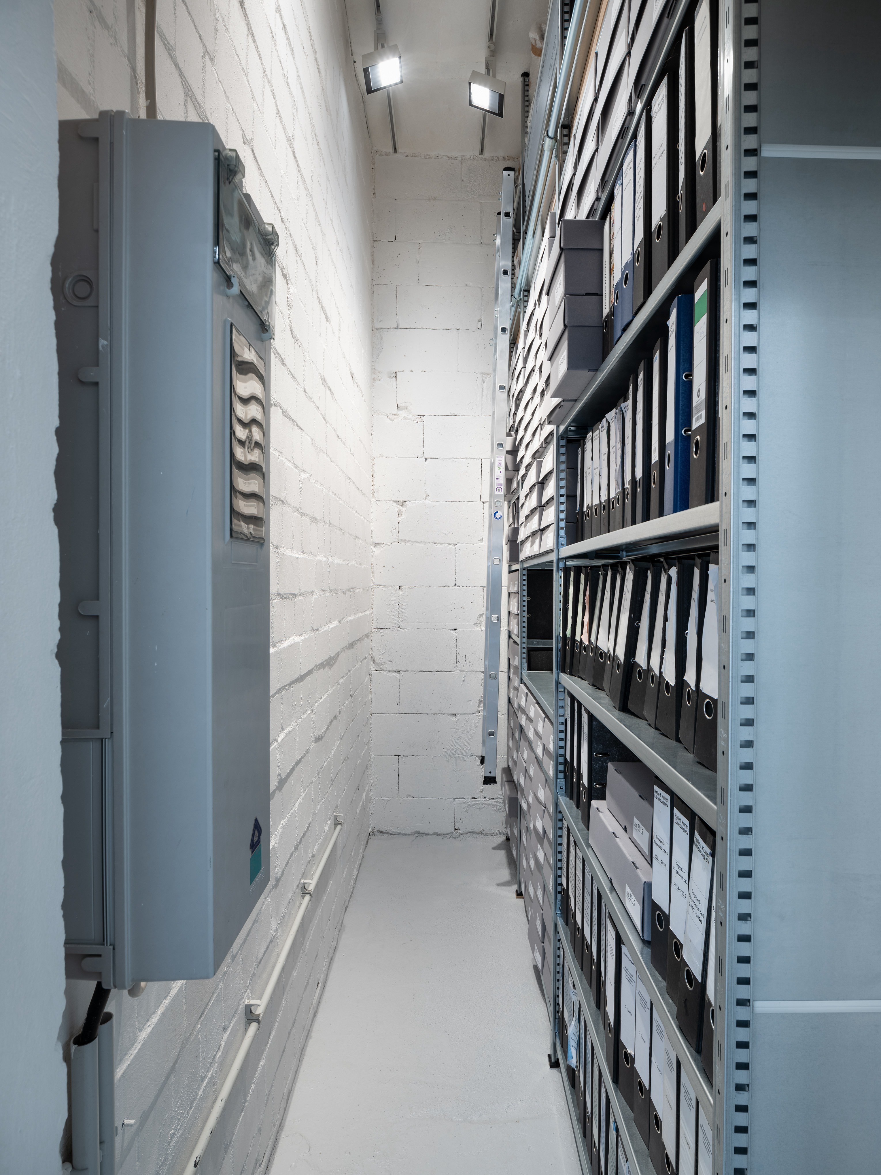 Archiv / Archive, Shelving system, lighting system, shelf ladder, wall paint, floor paint, archive boxes with adhesive labels, vinyl lettering, 350x113x391cm, 2025, Photography: Fred Dott
